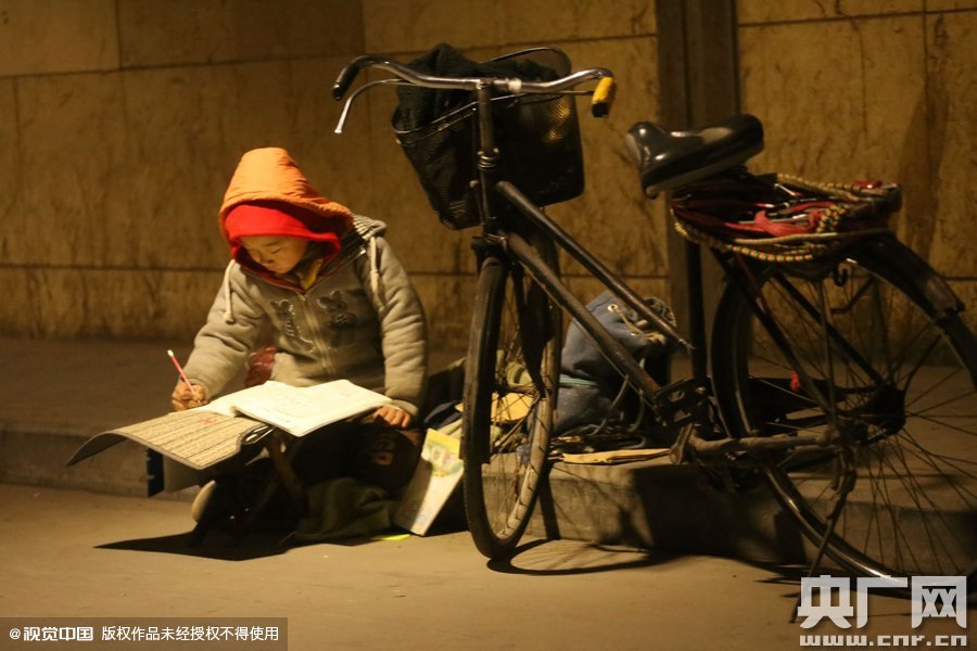 鄭州中原路京廣鐵路大橋下涵洞的馬路牙子邊，一位戴著紅色辣椒帽的小男孩趴在一個(gè)小馬扎上，上面鋪著一張車(chē)墊，在微弱的燈光下寫(xiě)作業(yè)