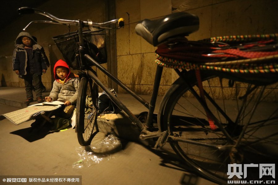 2015年11月8日晚，鄭州，7歲男孩涵洞橋下陪父母練攤，路邊燈下坐小馬扎上寫(xiě)作業(yè)。